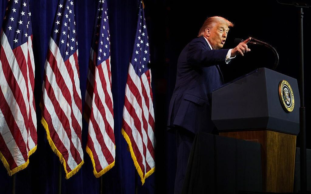 तस्वीर में अमेरिकी राष्ट्रपति डोनाल्ड ट्रम्प वॉशिंगटन में एक सार्वजनिक कार्यक्रम के दौरान मंच से संबोधित करते नजर आ रहे हैं।