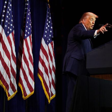 तस्वीर में अमेरिकी राष्ट्रपति डोनाल्ड ट्रम्प वॉशिंगटन में एक सार्वजनिक कार्यक्रम के दौरान मंच से संबोधित करते नजर आ रहे हैं।