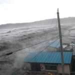 समुद्र किनारे ऊंची लहरें उठती हुई दिखाई दे रही हैं, तटीय इलाकों में चेतावनी के संकेत मौजूद हैं और लोग सुरक्षित स्थानों की ओर जा रहे हैं।