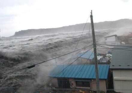 समुद्र किनारे ऊंची लहरें उठती हुई दिखाई दे रही हैं, तटीय इलाकों में चेतावनी के संकेत मौजूद हैं और लोग सुरक्षित स्थानों की ओर जा रहे हैं।