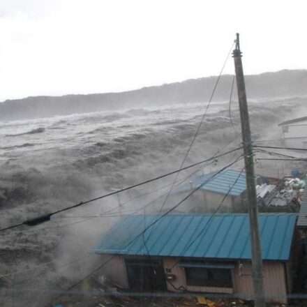 समुद्र किनारे ऊंची लहरें उठती हुई दिखाई दे रही हैं, तटीय इलाकों में चेतावनी के संकेत मौजूद हैं और लोग सुरक्षित स्थानों की ओर जा रहे हैं।